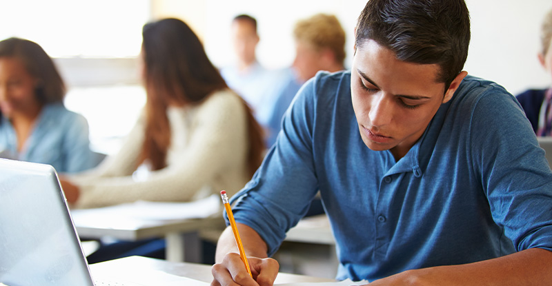 A photo of students seated in a classroom with the focus placed on a boy writing in a notebook with his laptop open.